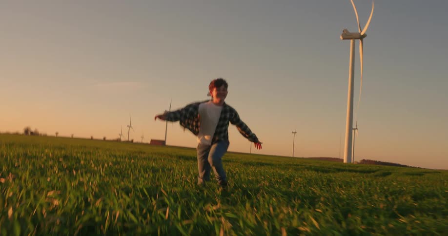 A boy smiles while running through a wind farm at sunset