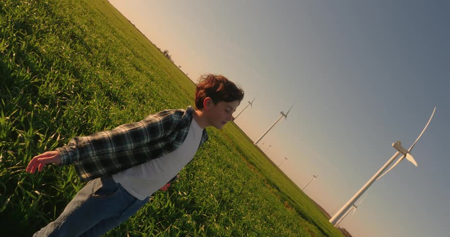 With his arms outstretched and eyes closed a young boy stands still on a flat grassland feeling the wind blowing around him amidst a field of wind turbines