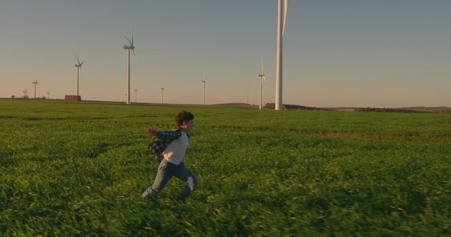 Boy running through a green field with wind energy platforms