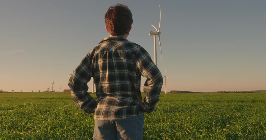 A boy stands on a wind farm and looks up at a wind mill on a windy day