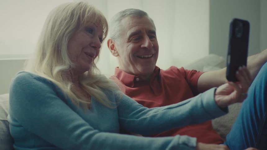 A man and a woman with gray hair are sitting on a sofa looking at a cell phone and are smiling while talking