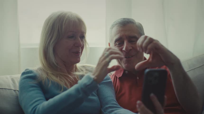 A couple sits on a couch in a bright room near a window smiling and making heart shapes with their hands while engaged in a video call