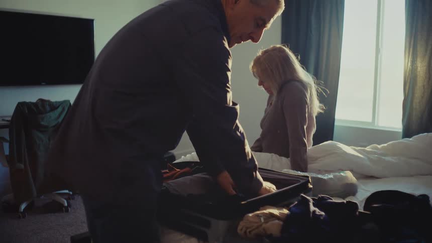 A man packs clothes into a suitcase on the bed while a woman sits nearby bathed in sunlight streaming through the window