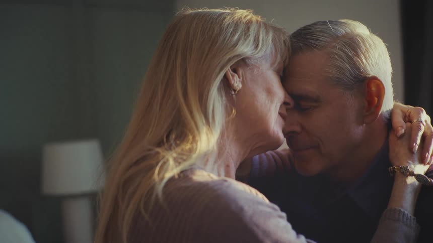 A mature couple shares an intimate and tender moment as a woman with long blonde hair gently holds the head of an older man with short gray hair