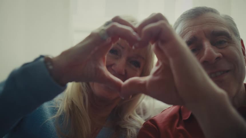 A couple sits on a couch in a bright room near a window smiling waving goodbye and making heart shapes with their hands while engaged in a video call