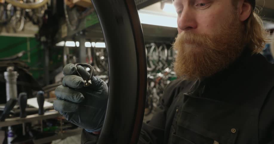 A bearded man wearing a beanie and gloves inspects the valve stem area of a bicycle wheel in a workshop