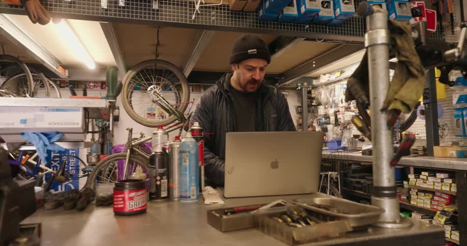 A mechanic is using a laptop to research or troubleshoot while surrounded by tools and bicycle parts in his workshop