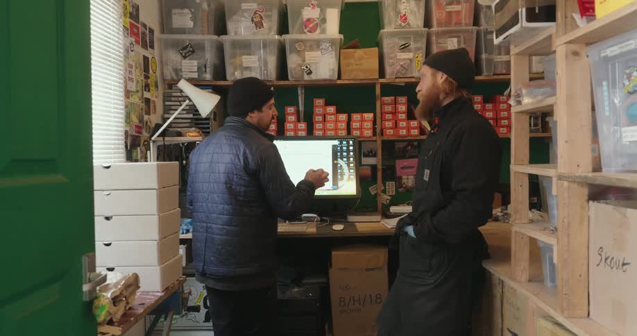 Two men are talking in a cluttered workshop or stockroom one standing at a computer desk and the other leaning against it