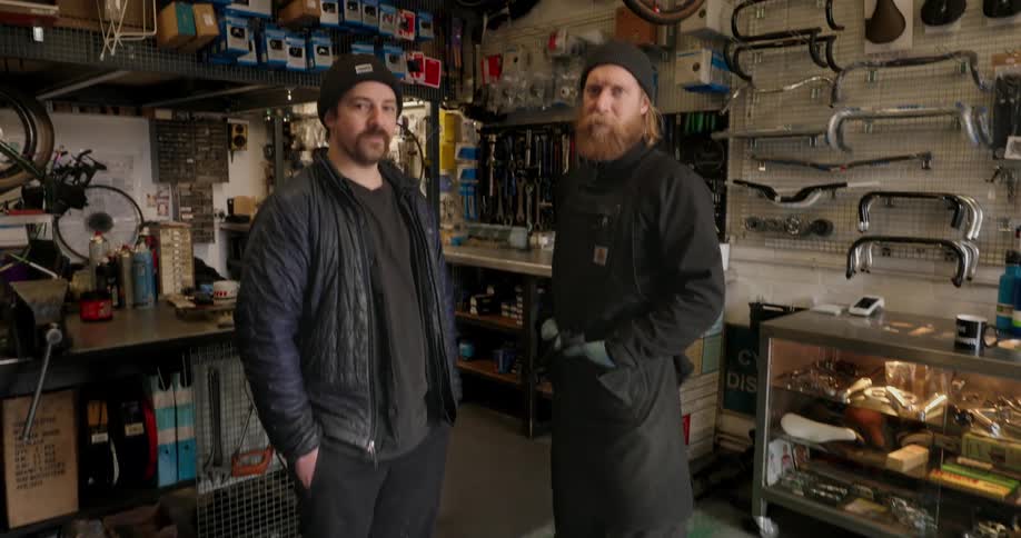 Two men stand looking at the camera inside a cluttered bicycle repair shop filled with parts tools and wheels