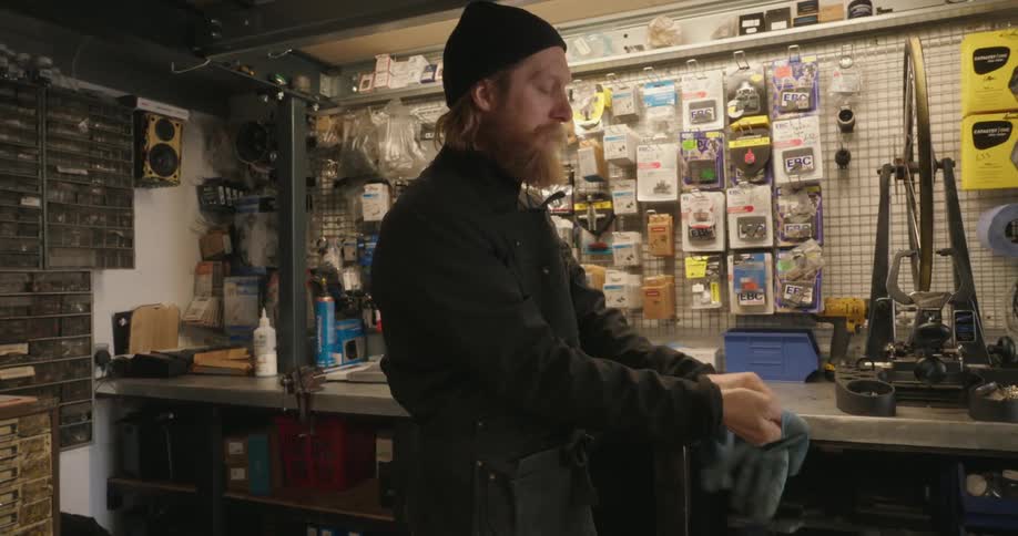 A man in a black cap wearing a jacket is blowing on a blue glove to put it on his hand in a workshop