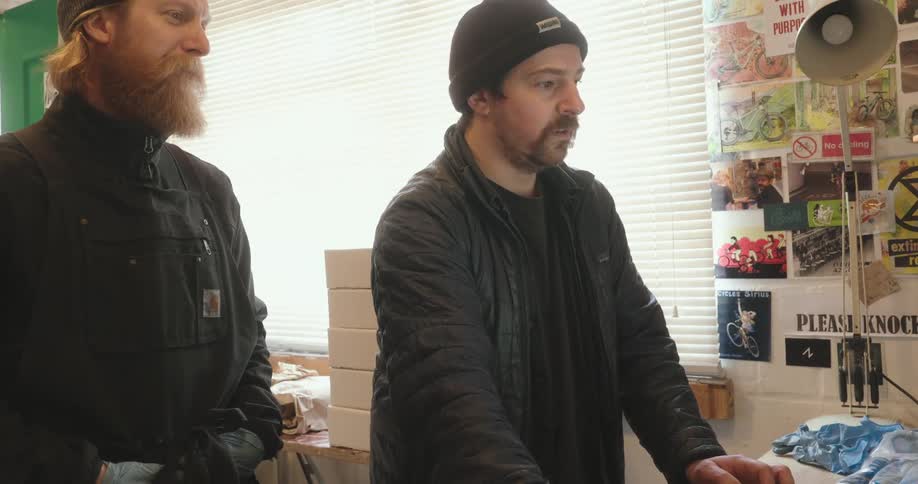 Two men stand side by side in a cluttered workshop discussing something off screen