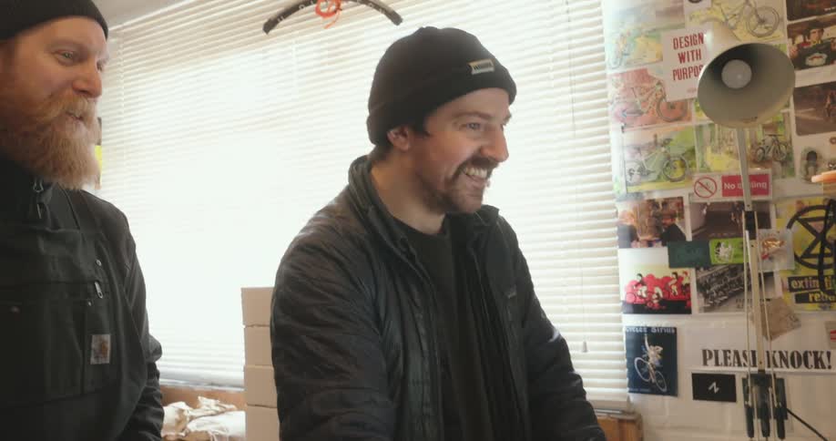 Two men wearing beanies smile while having a conversation in a cluttered workshop office