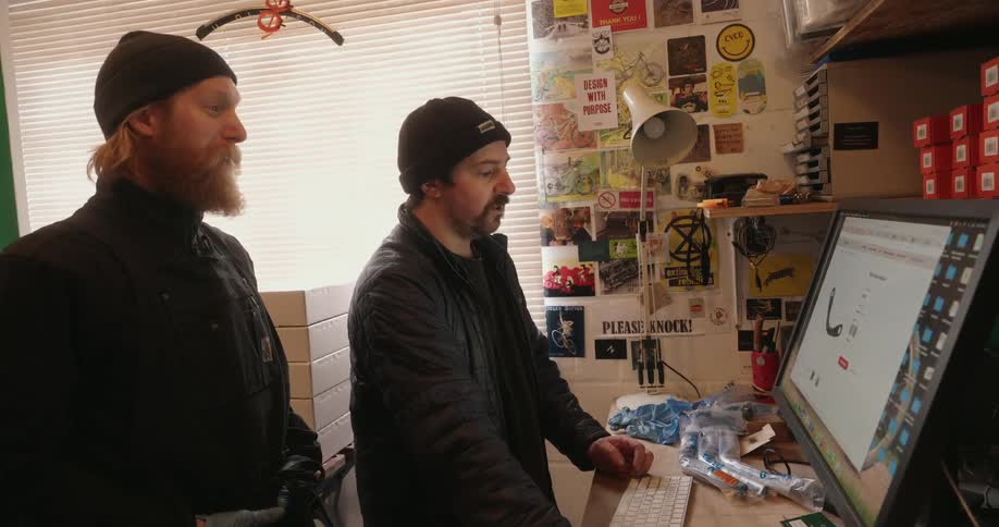 Two bearded men intently look at a computer screen together in a messy creative workspace filled with posters and equipment