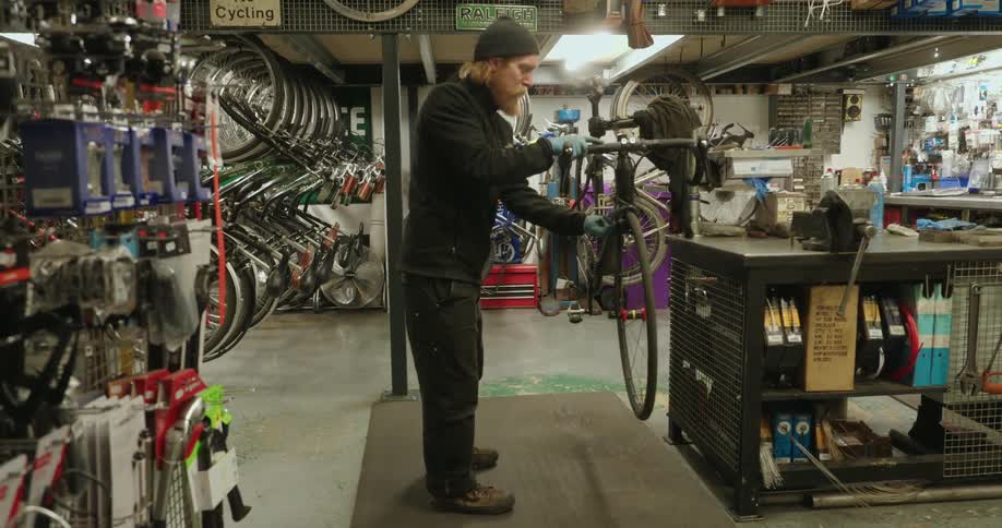 A cluttered workshop with a person repairing a bicycle surrounded by scattered tools and spare parts