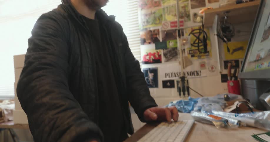 Standing at a cluttered desk in a workshop office a man uses a mouse then types on an imac computer while talking to another man partially offscreen as the camera pans up