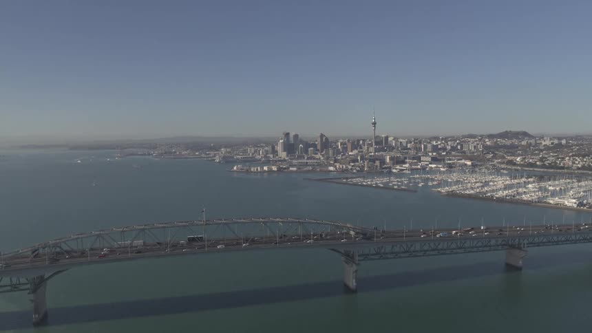 An aerial view shows aucklands cityscape and harbor under a clear sky with a trafficladen bridge spanning the foreground