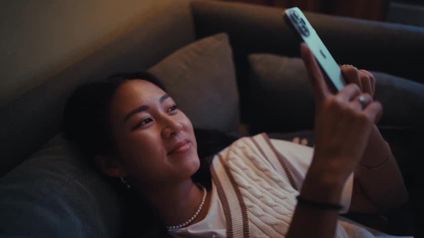 Woman reclining on her couch smiles while using her smartphone for a video call