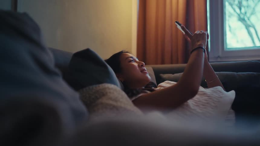 Woman lying on her couch smiles while using her smartphone for a video call