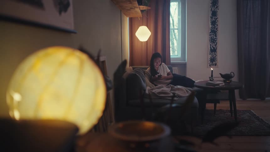 A woman reclines on a couch in a dimly lit room engrossed in a smartphone with lamps glowing nearby and a candle on the table