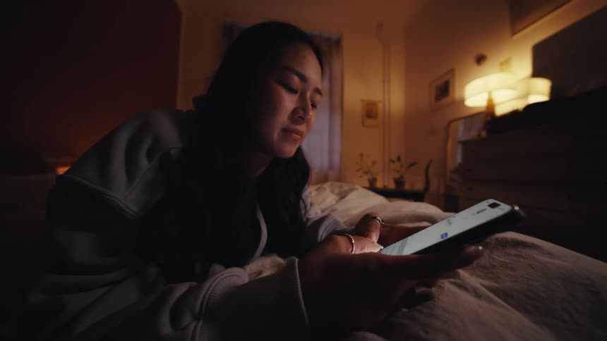 Young woman lying on her bed in a dimly lit bedroom texts on her smartphone