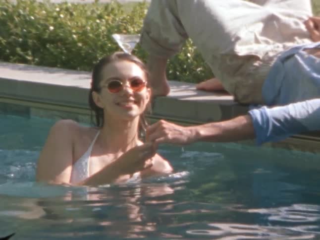 Two people are relaxing by a swimming pool with one person in the water and the other lying on the edge reaching out to hold hands
