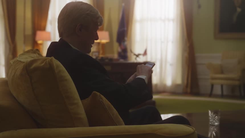 A man with gray hair and a black suit sitting on a yellow sofa using his cell phone