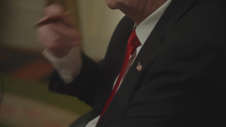 The torso of a man wearing a black suit with a red tie and a brooch with a country flag