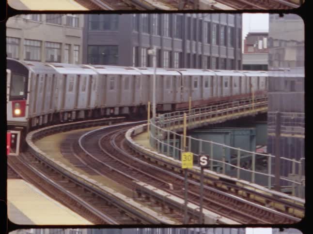A subway train approaches a platform on a curved track framed by urban buildings and a person waiting nearby