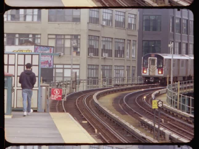 A subway train approaches a platform on a curved track framed by urban buildings in the background and a person waiting nearby