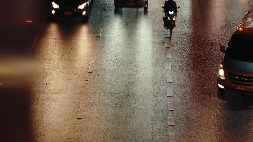 A series of cars motorcycles and a bus driving along a busy road at night with their lights reflecting on the road