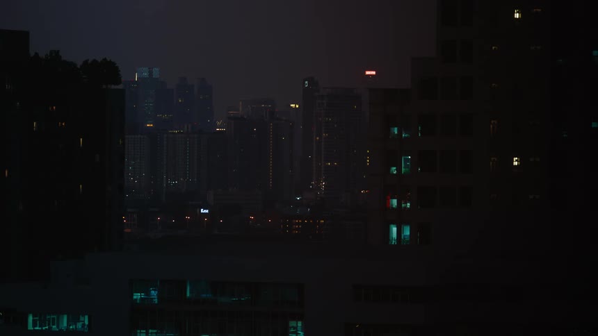A city view with a series of skyscrapers faintly reflecting light from the buildings as the environment remains dark due to nighttime