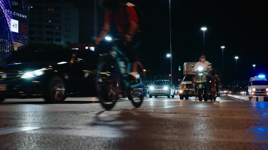 Cars motorcycles and a tuk tuk drive along a busy wet looking city street illuminated by headlights and streetlights at night