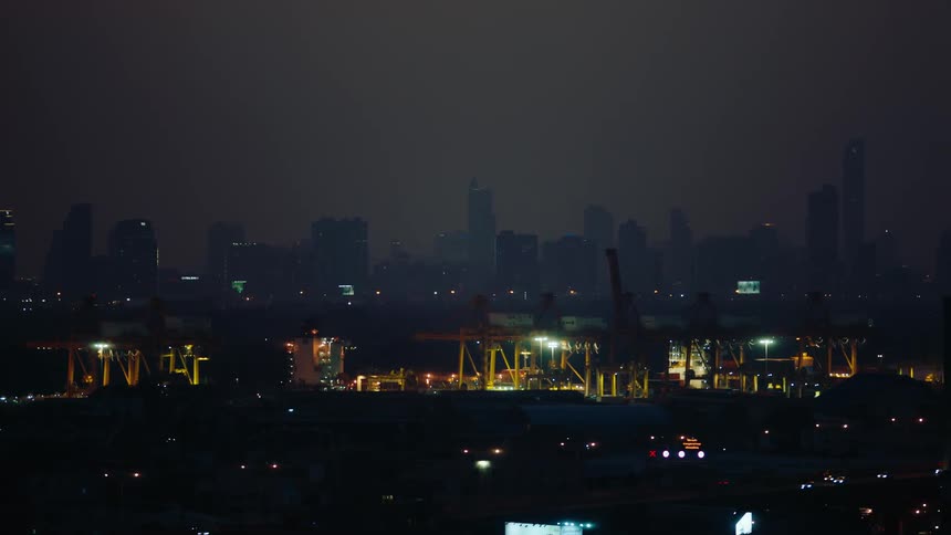 Traffic drives along a highway at night near a large construction site while fog covers the skyline in the background
