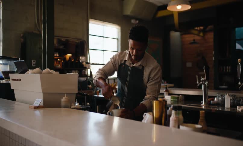 Male barista pours hot milk into a cup in an empty coffee shop during the day