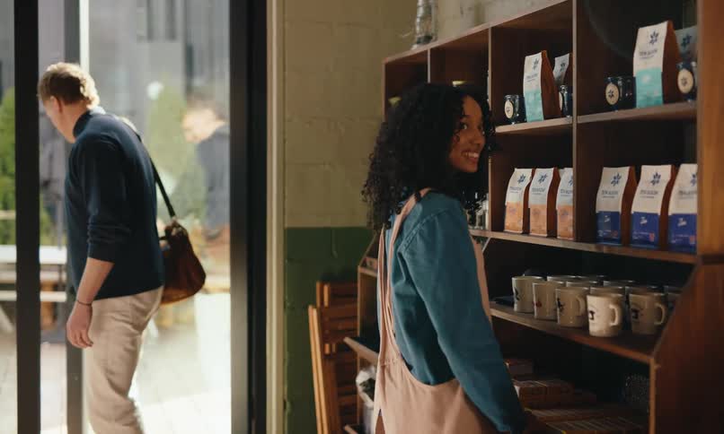Man walking into a coffee shop and a woman arranging mugs on a shelf