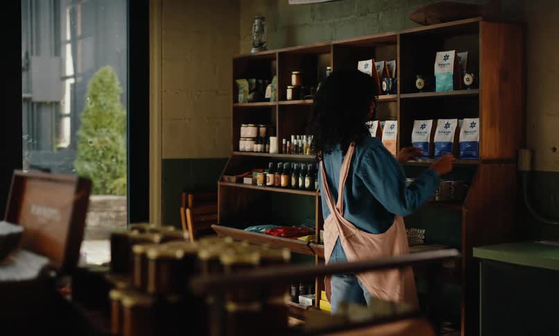 A woman wearing a blue shirt and apron arranges items on shelves stocked with coffee bags mugs and decor in a welllit store