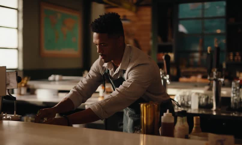 A bartender works behind a counter surrounded by various bottles and barware with sunlight streaming through a window