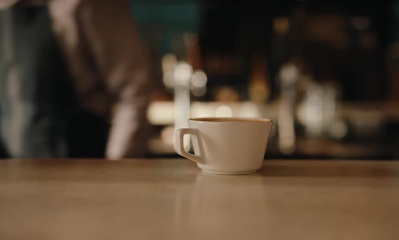 A barista partially obscured and wearing an apron gently places a white cup on a countertop