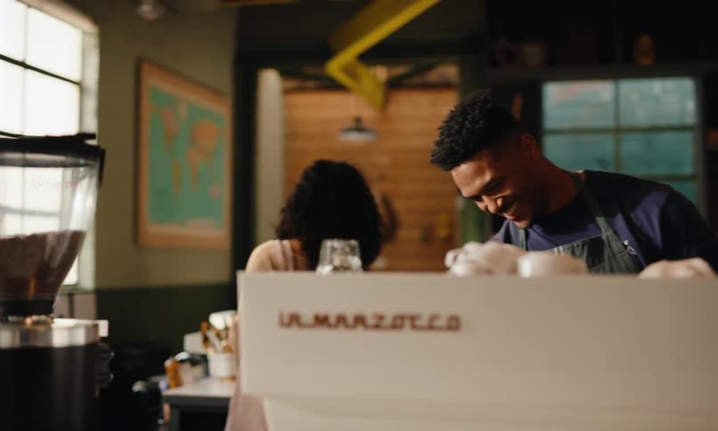 A young black man wearing an apron in a coffee shop is happily preparing coffee for someone smiling as a young woman walks behind him inside the shop
