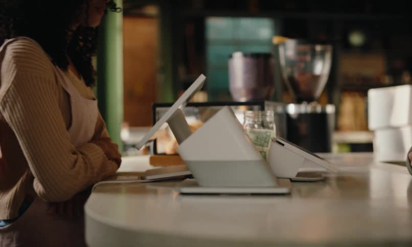 A woman taking another womans order with a tablet in a coffee shop walks over to a man wearing an apron and they both talk and smile at midday