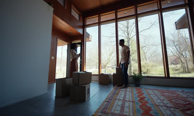 A couple hugs in a modern living room with large windows surrounded by moving boxes and a colorful rug