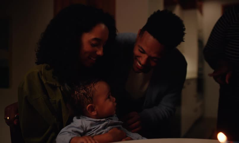A father mother and grandmother sing happy birthday to a baby sitting on the mothers lap with a cake holding a single candle in a dimly lit room