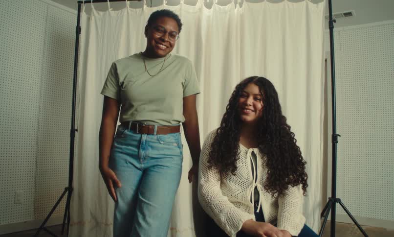 Two women are positioned in front of a white curtain backdrop with one standing and the other sitting on a chair in a room equipped with photography lighting equipment