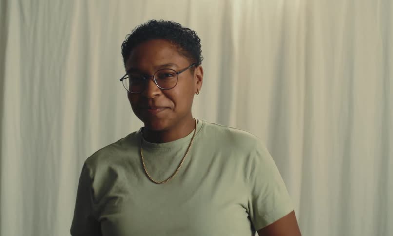 A person in a light colored shirt smiles at the camera while adjusting their glasses with both hands against a plain curtain backdrop