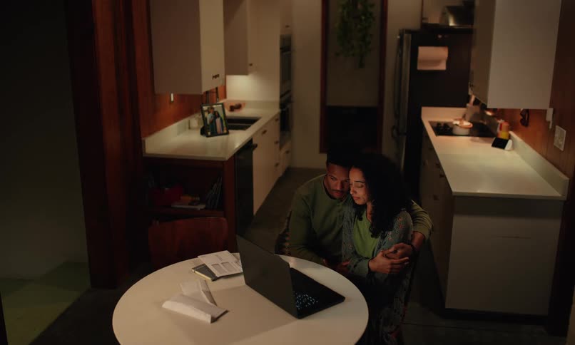 A man walks over and hugs a woman by the laptop
