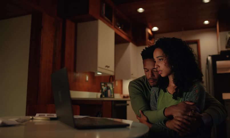 A man warmly embraces his partner consoling her as they focus on a laptop in the soft warm light of the room