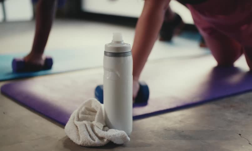 Two women are exercising with dumbbells on a yoga mat, and one of them quickly grabs a white water bottle beside her to take a drink.