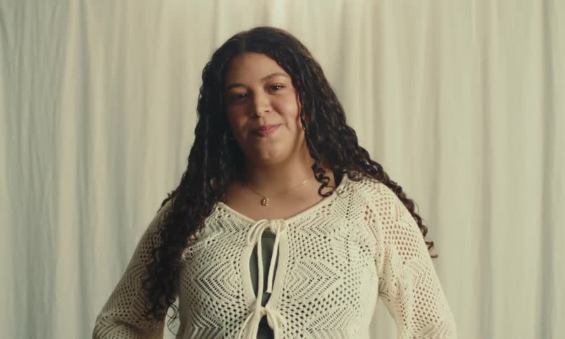 A woman stands in front of a lightcolored curtain wearing a white crocheted top with a tiefront detail