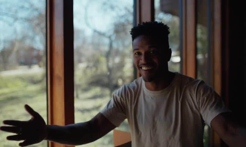 A young happy man places a moving box down turns around and greets someone in a sunlit room with large windows