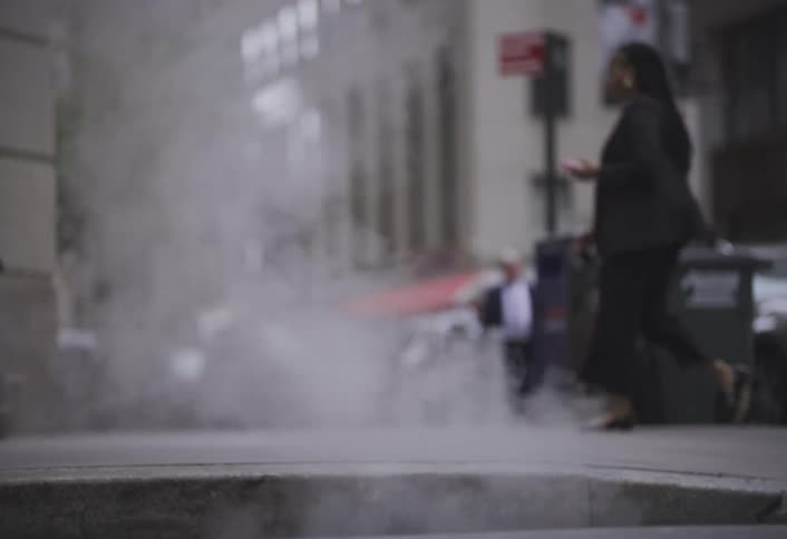 Steam rises from a manhole cover as several people casually walk by in a bustling city scene